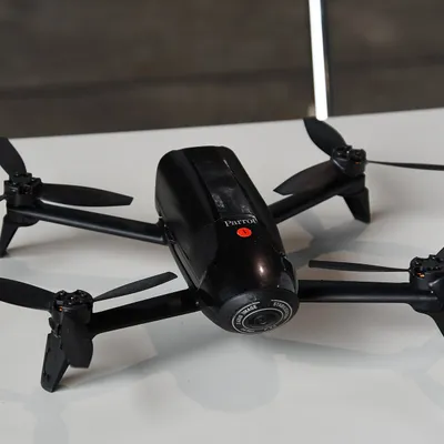 A black Parrot Bebop 2 on a white table, seen from the front left, with its fisheye camera pointing at the viewer.